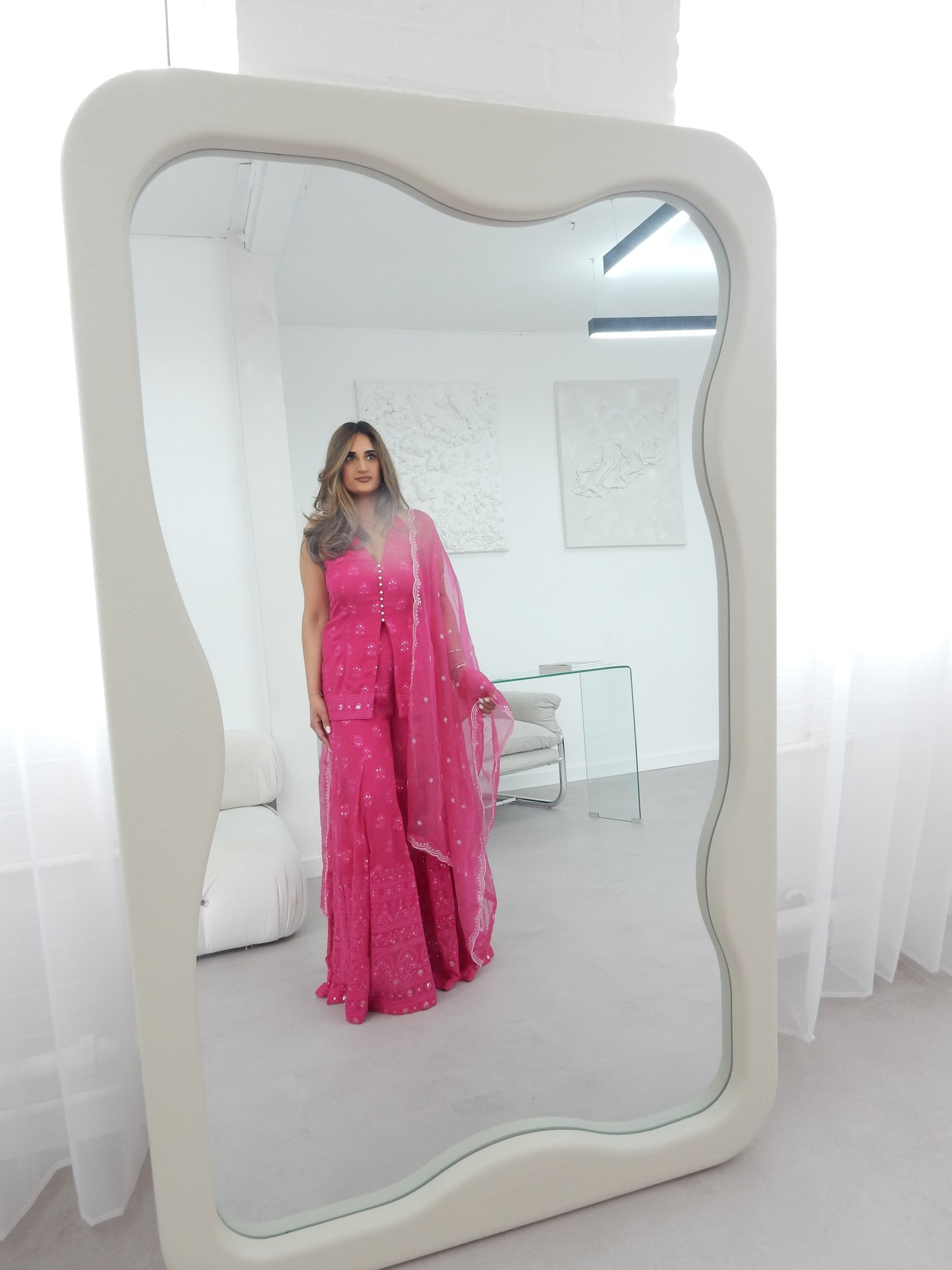 Indian Woman in a halter neck hot pink shararah standing in front of a large mirror. The background shows a white background with white modern furniture. Ideal for a jaggo or Mendhi event.