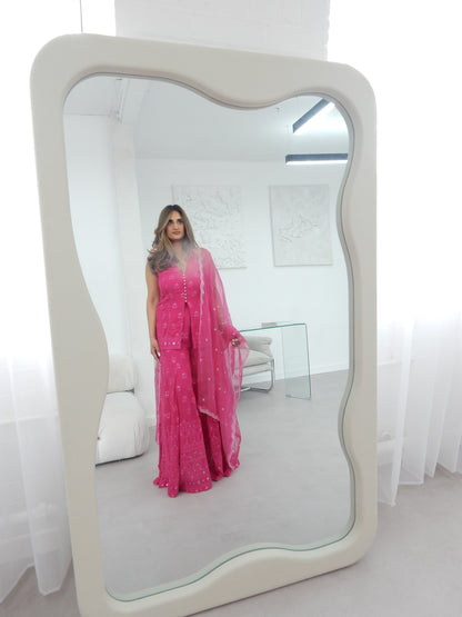 Indian Woman in a halter neck hot pink shararah standing in front of a large mirror. The background shows a white background with white modern furniture. Ideal for a jaggo or Mendhi event.