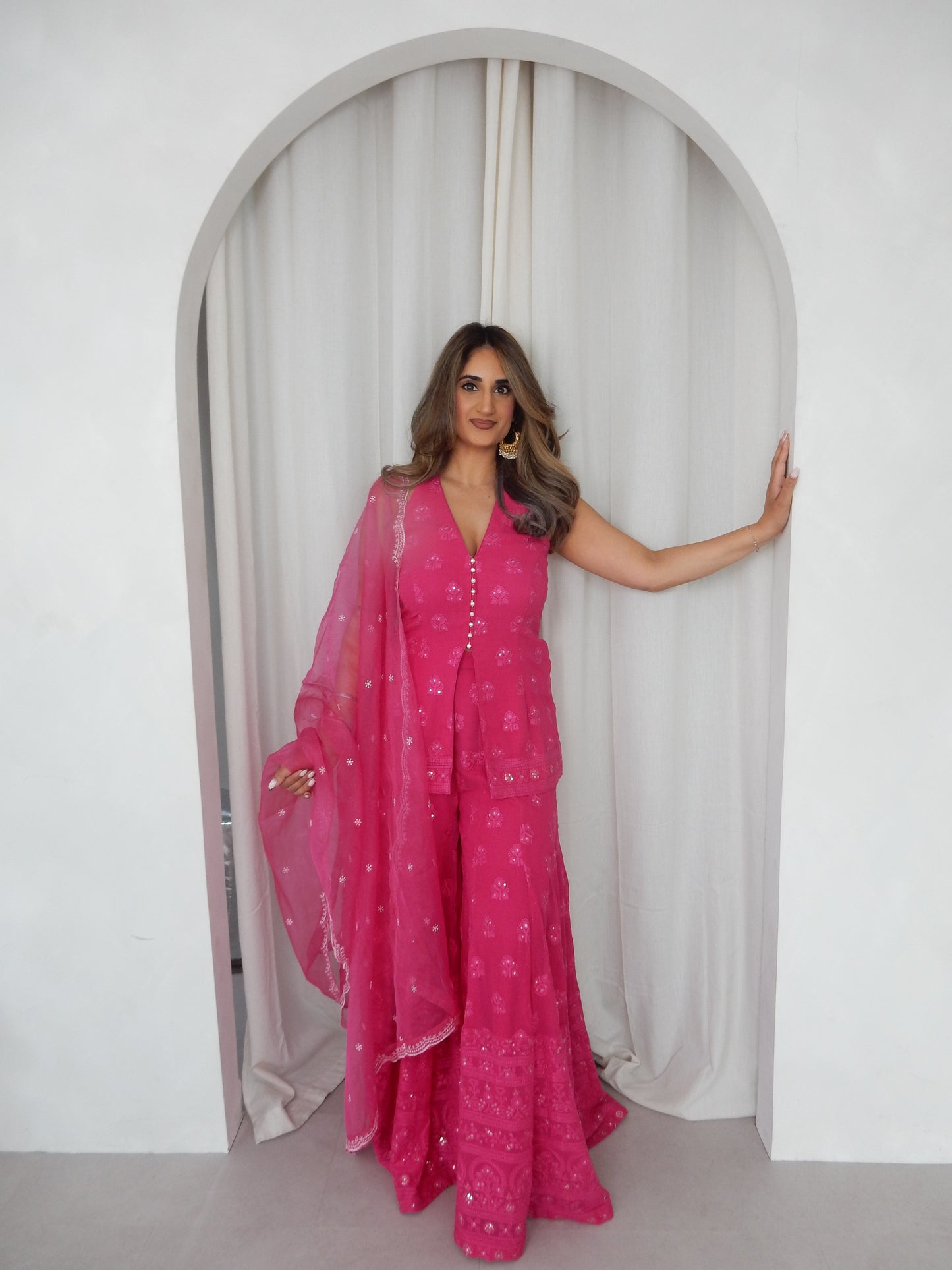 Indian Woman in a halter neck hot pink shararah standing behind a white arch way. Ideal for a jaggo or Mendhi event.