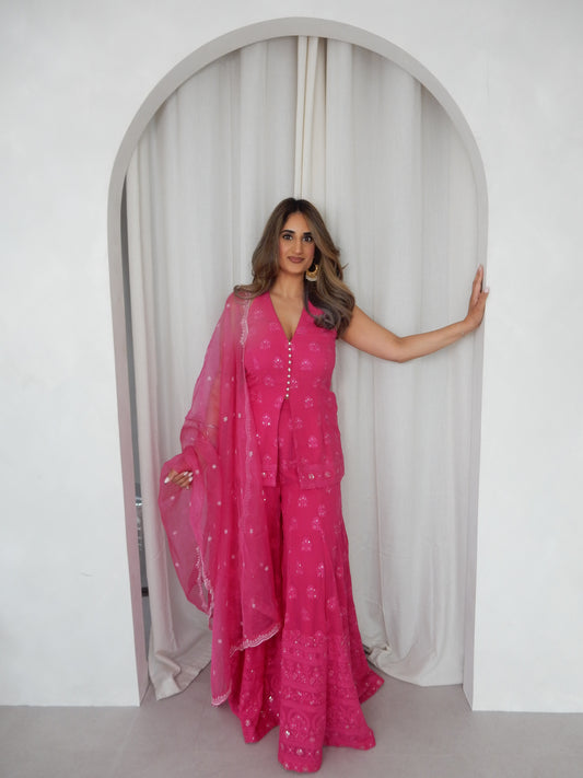 Indian Woman in a halter neck hot pink shararah standing behind a white arch way. Ideal for a jaggo or Mendhi event.