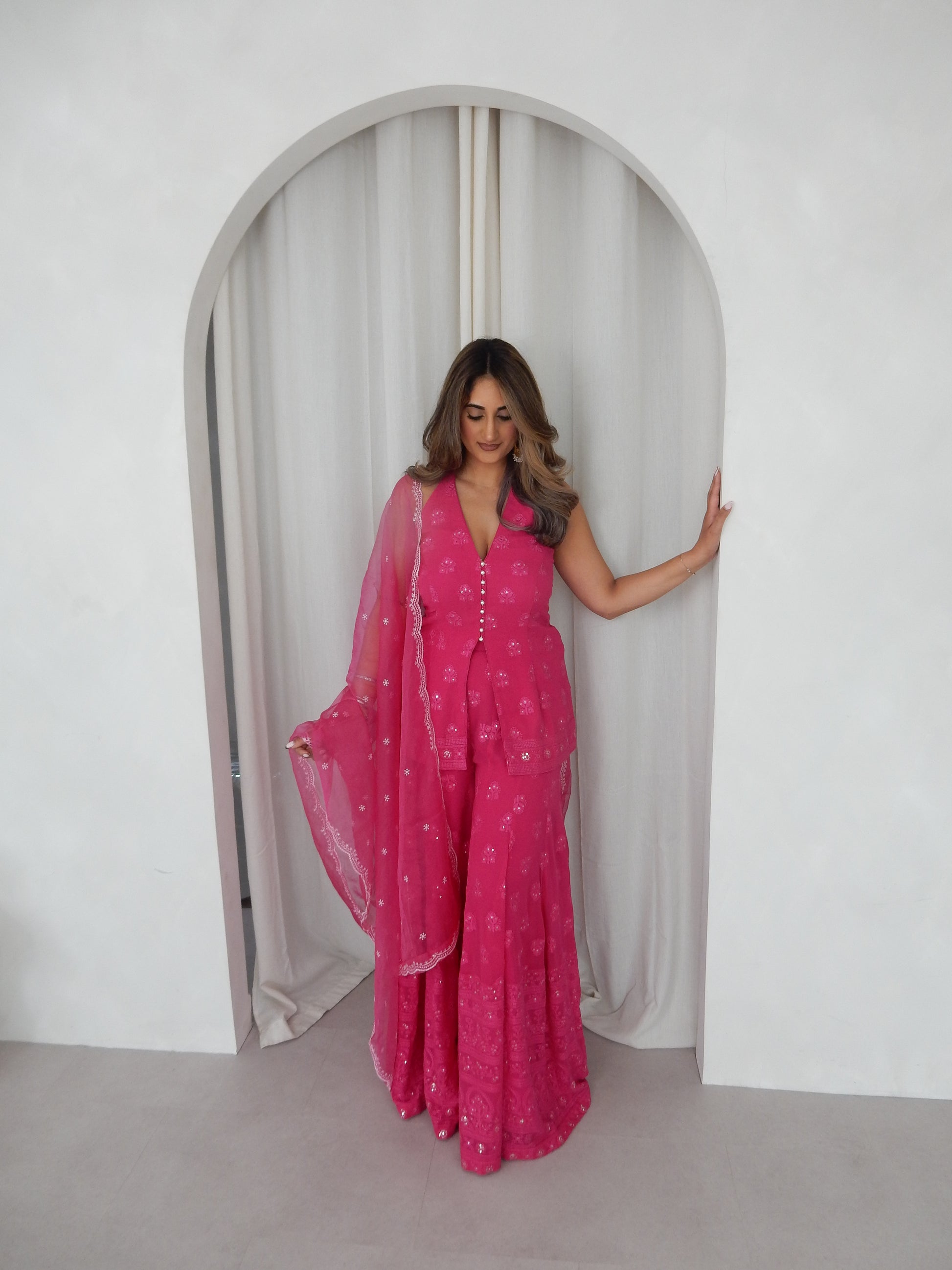 Indian Woman in a halter neck hot pink shararah standing behind a white arch way. Ideal for a jaggo or Mendhi event.