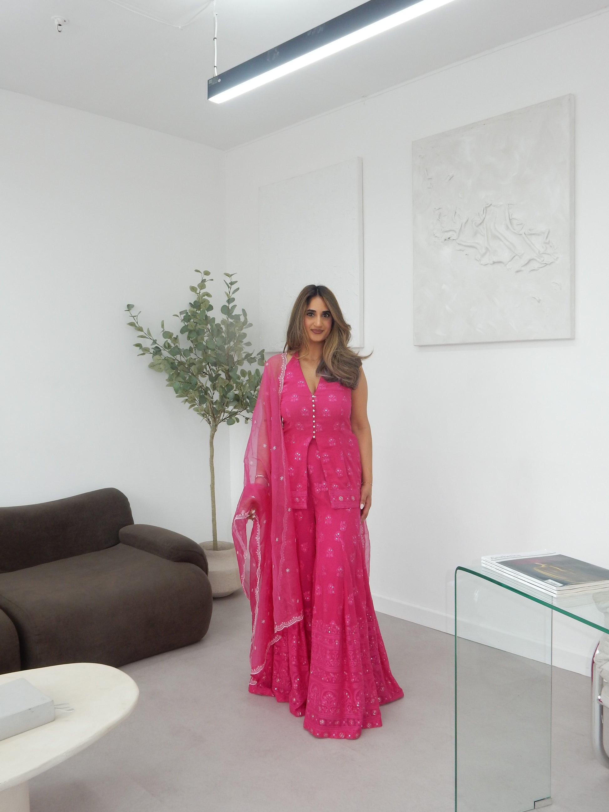 Indian Woman in a halter neck hot pink shararah standing in a room with a white wall and brown sofa. Ideal for a jaggo or Mendhi event.