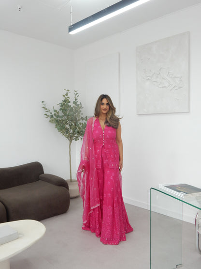 Indian Woman in a halter neck hot pink shararah standing in a room with a white wall and brown sofa. Ideal for a jaggo or Mendhi event.