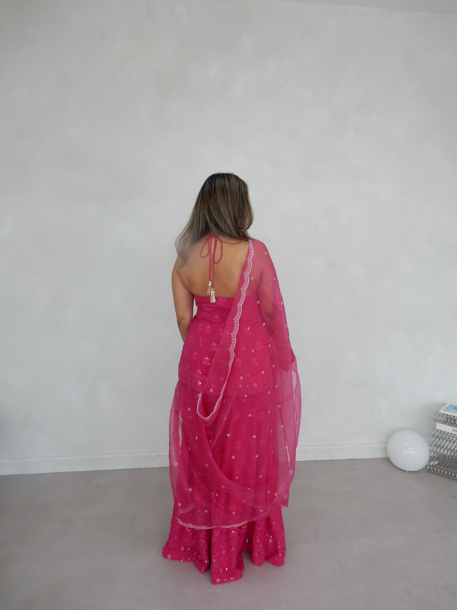 Indian Woman in a halter neck hot pink shararah facing towards the wall as her back is displayed. She is standing in front of a light grey background. Ideal for a jaggo or Mendhi event.